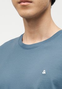 Νεαρός άνδρας που φοράει μπλε t-shirt με στρογγυλή λαιμόκοψη και μικρό λευκό λογότυπο αμφικρανού (&) στο αριστερό στήθος, σε απλό λευκό φόντο.