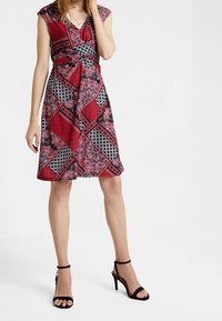 Rotes Kleid mit kurzen Ärmelchen, das ein geometrisches Muster in Schwarz und Weiß zeigt. Ein geknotetes Taillen-Design und ein fließender Rock. Schwarze Riemchenabsätze.