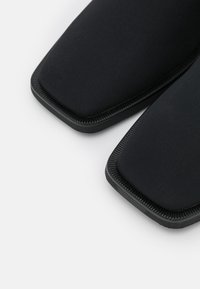 Zapatos negros de tipo slip-on que presentan una parte superior de textil suave con una puntera cuadrada y una suela de goma texturizada. Diseño minimalista sin acentos visibles.