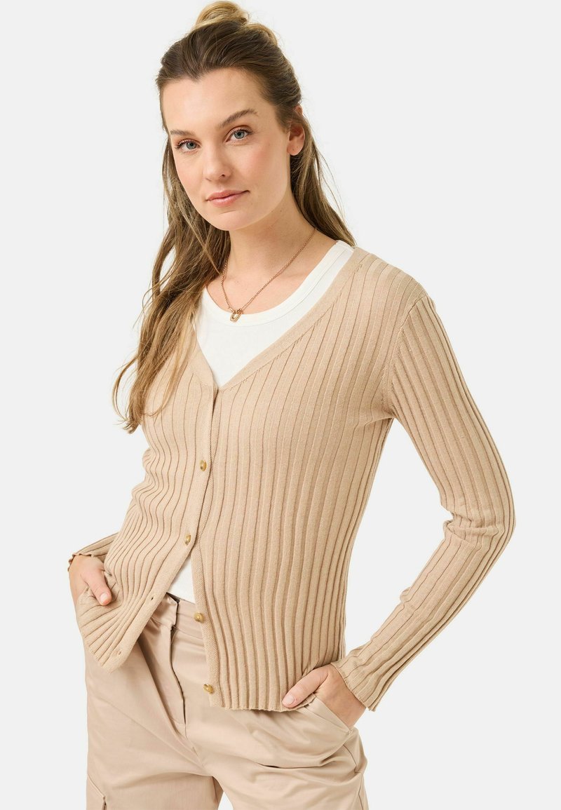Beige gerippter Cardigan mit tiefem V-Ausschnitt und vorderen Knöpfen. Lange Ärmel, leichtes Design, kombiniert mit einem weißen Oberteil und lässigen Hosen.