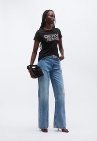 Svart t-skjorte med "DKNY JEANS"-logo i rhinsten, kombinert med lyseblå vide jeans. Holder en svart håndveske, iført røde sandaler med hæl.