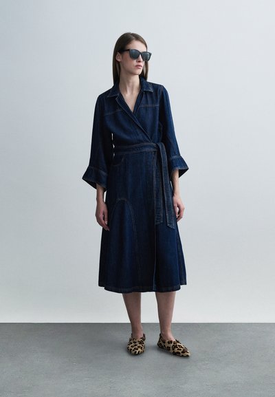 Robe portefeuille en jean de couleur bleu profond, manches longues avec poignets évasés, et ceinture à nouer à la taille. Portée avec des chaussures plates à imprimé animal.