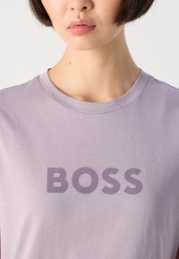 Aukštas asmens liemens vaizdas, turinčio trumpus plaukus, vilkintis šviesiai purpurinę marškinėlį su žodžiu "BOSS", atspausdintu tamsiai purpurine spalva krūtinėje.