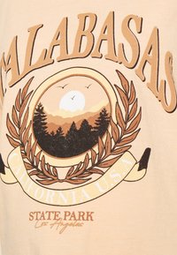 T-shirt bege com uma gráfica de um pôr do sol sobre montanhas e árvores, com "Calabasas California U.S.A" em tipografia em negrito.