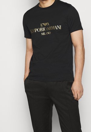 Homme portant un t-shirt noir Emporio Armani Milano avec un logo doré, associé à un pantalon noir, debout avec une main dans la poche.