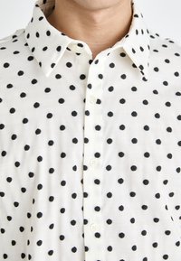Biela košeľa s gombíkmi, na ktorej sa nachádza vzor rovnomerne rozmiestnených čiernych bodiek. Tkanina vyzerá ľahká s hladkou textúrou.