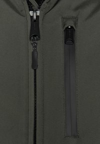 Veste vert olive avec une texture lisse, dotée d'une fermeture éclair centrale avec tirette noire et d'une poche zippée sur le côté pour le rangement.