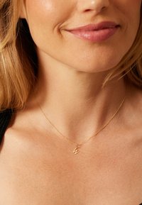 Goldkette mit einer dünnen Kette und einem kleinen "W"-Anhänger, getragen um den Hals auf glatter, gebräunter Haut.