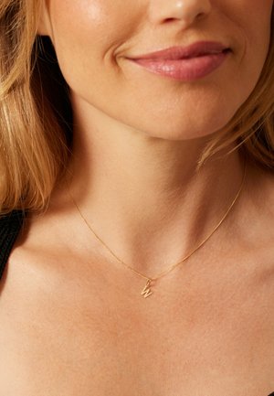 Collier en or avec une chaîne fine et un petit pendentif en forme de 'W', porté autour du cou sur une peau lisse et bronzée.