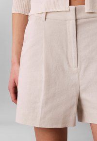 Primer plano de una persona que lleva pantalones cortos beige con textura y un suéter acortado de canalé, de pie frente a un fondo gris liso.