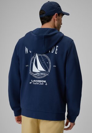 Mężczyzna w granatowej bluzie z kapturem i czapce, na bluzie jest biały żaglówka i tekst "Lakeside Yacht Club" na plecach.