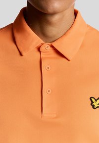 Kortærmet orange polo skjorte lavet af struktureret stof, med tre knapper og en sort ørne logo på venstre bryst.