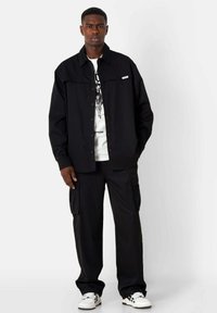 Zwarte button-up shirt met een relaxte pasvorm, witte graphic t-shirt eronder, gecombineerd met zwarte cargobroek en witte sneakers met zwarte accenten.