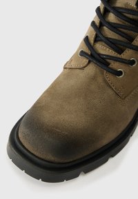 Bota cano curto em camurça castanha com atacadores pretos, bico redondo, parte superior texturizada e sola de borracha preta grossa com padrão de tração.