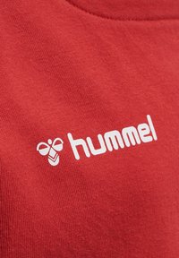 Tessuto di cotone rosso con un logo bianco che presenta un'illustrazione di un'ape e la parola "hummel" in un carattere bold e moderno. Texture liscia, design informale.