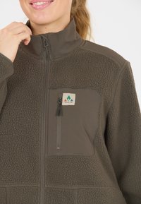 Giacca in pile marrone con colletto liscio, tasca con zip e tessuto testurizzato. Presenta un patch del logo sul petto con colori a contrasto.