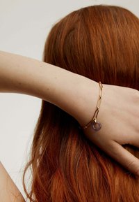 Una mano con una pulsera de cadena de oro que presenta un charm circular púrpura reposa sobre cabello rojo largo, contra un fondo neutro.