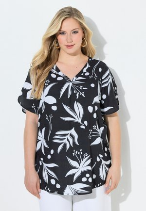 Frau mit langen blonden Haaren, die goldene Creolen und ein schwarzes kurzärmeliges Oberteil mit weißem botanischem Druck trägt, steht vor einem weißen Hintergrund.