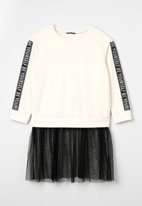 Top de manga larga crema con la inscripción "SE TÚ MISMO" en letras negras en las mangas, combinado con una falda de tul negra. Tela suave y diseño con capas.