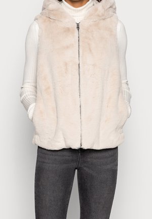 Kobieta ubrana w bezrękawnik z beżowego sztucznego futra na zamek z kapturem, biały prążkowany golf oraz ciemnoszare dżinsy, ręce w kieszeniach.