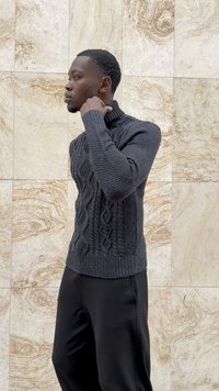Mann in einem dunkelgrauen, grob gestrickten Rollkragenpullover und schwarzen Hosen, der seitlich blickt, während er vor einer beigefarbenen, strukturierten Steinwand steht.