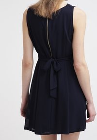 Osoba otočená zády, oblečená v bezrukávových námořnických šatech s uvázaným pasem a viditelným zipem vzadu, na neutrálním pozadí.