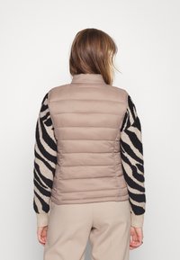 Prošívaná vesta světle béžové barvy s vysokým límcem a černo-bílými proužkovanými pletenými rukávy. Má přiléhavý střih a žebrované manžety.