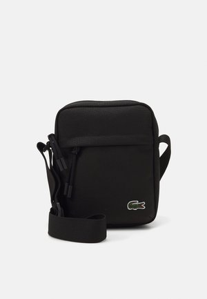 Sort kompakt crossbody-taske med justerbar rem, lynlåslomme foran og grønt krokodillelogo i nederste højre hjørne.