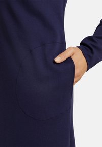 Marinblå klänning med rund ficka och långa ärmar, tillverkad av ett slätt tyg. Fickdetaljen kontrasterar med materialets textur.