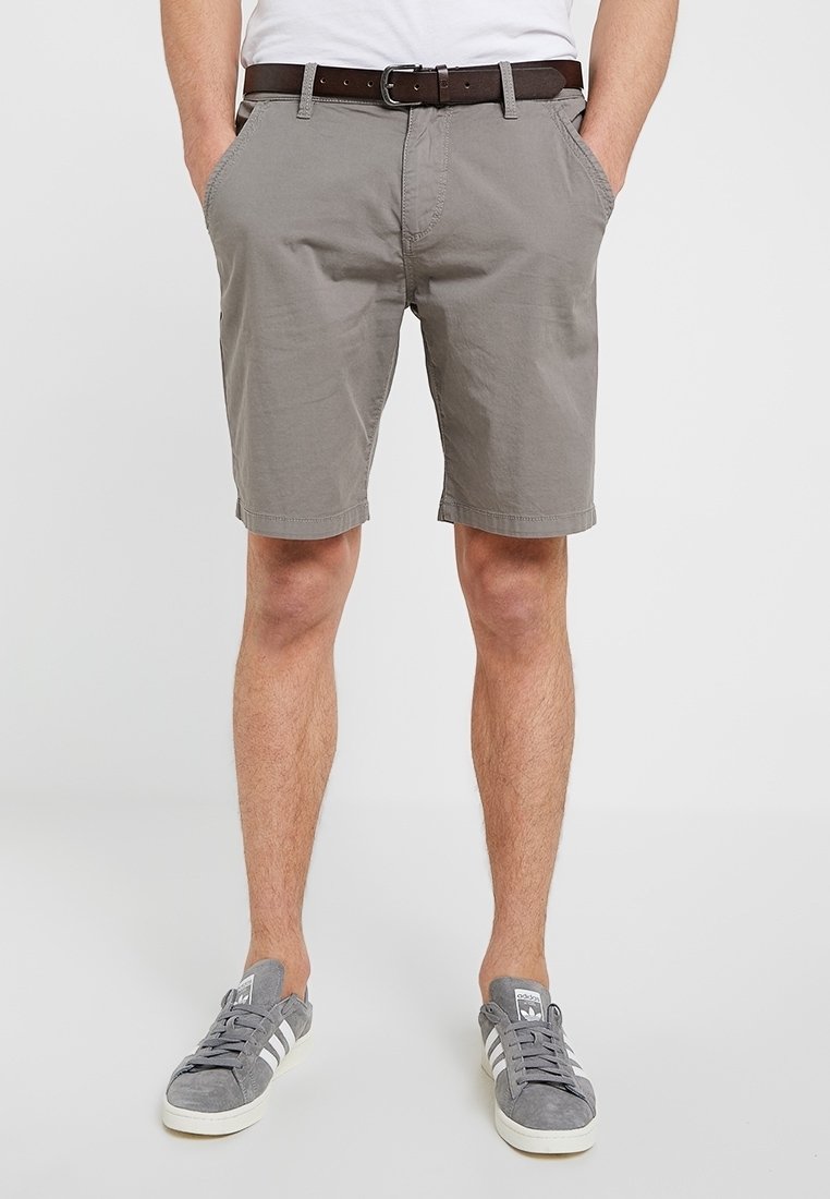 Homme portant un short gris mi-long avec poches latérales, une ceinture marron et des baskets Adidas grises avec des bandes blanches.