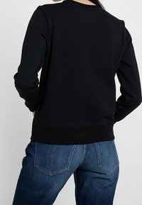 Czarna bluza z okrągłym dekoltem, ściągaczami przy rękawach i na dole. W połączeniu z niebieskimi dżinsami z zaokrąglonymi kieszeniami z tyłu i luźnym fasonem.