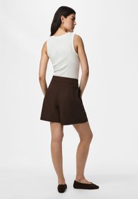 Femme debout de côté, mains dans les poches, portant un haut blanc sans manches, un short marron foncé taille haute et des chaussures plates foncées.