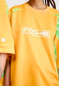 Tricou sportiv portocaliu cu mâneci scurte, logo alb și accente din plasă verzi pe părțile laterale. Textură netedă cu detalii din cusătură verde.