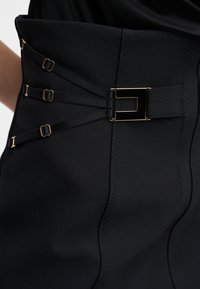 Κοντινή λήψη μαύρου ενδύματος με ψηλή μέση, χρυσή ορθογώνια αγκράφα και τρεις λεπτές ρυθμιζόμενες τιράντες στο πλάι.