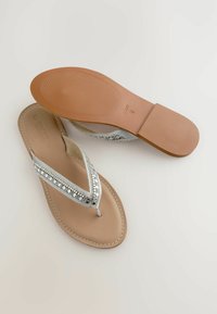 Silberne Flip-Flops mit einem bestickten Riemen, beigem Fußbett und Gummisohle. Die Bestickung zeigt schwarze und weiße Akzente mit kontrastierenden Nähten.