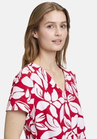 Rotes Kleid mit weißem Blumenmuster, kurzen Ärmeln, V-Ausschnitt und lockerer Passform. Glatte Stoffstruktur mit kräftigen, lebendigen Farben.