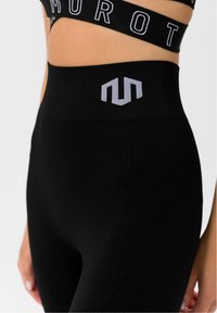 Czarne prążkowane legginsy mają wysoki pas z jasnoszarym logo. Materiał jest elastyczny i gładki, zapewniając dopasowany krój.