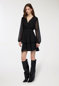 Vestito nero a pois con scollatura a V profonda, gonna a strati e maniche lunghe trasparenti. Abbinato a stivali neri lucidi sopra il ginocchio.