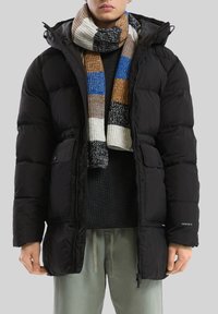 Čierna páperová bunda s kapucňou, štýlový dizajn, textúrovaná látka a predné vrecká. Viacfarebný pruhovaný pletený šál prehodený okolo krku.