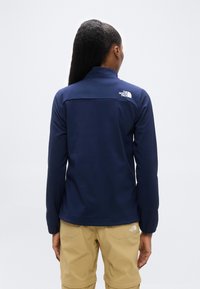 Άτομο με μακριά μαλλιά δεμένα σε κότσο, φοράει μπουφάν North Face σε σκούρο μπλε και μπεζ παντελόνι, φαίνεται από πίσω με φόντο απλό.