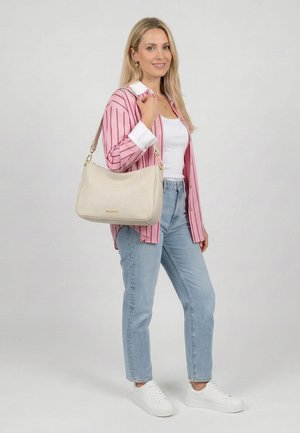 Frau trägt ein rosa gestreiftes Hemd, weißes Top, hellblaue Jeans, weiße Sneakers und trägt eine beige Umhängetasche, steht vor einem einfarbigen Hintergrund.