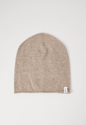 Beige knitted beanie with a small white fabric tag featuring black text on the bottom edge, laid flat on a white background.