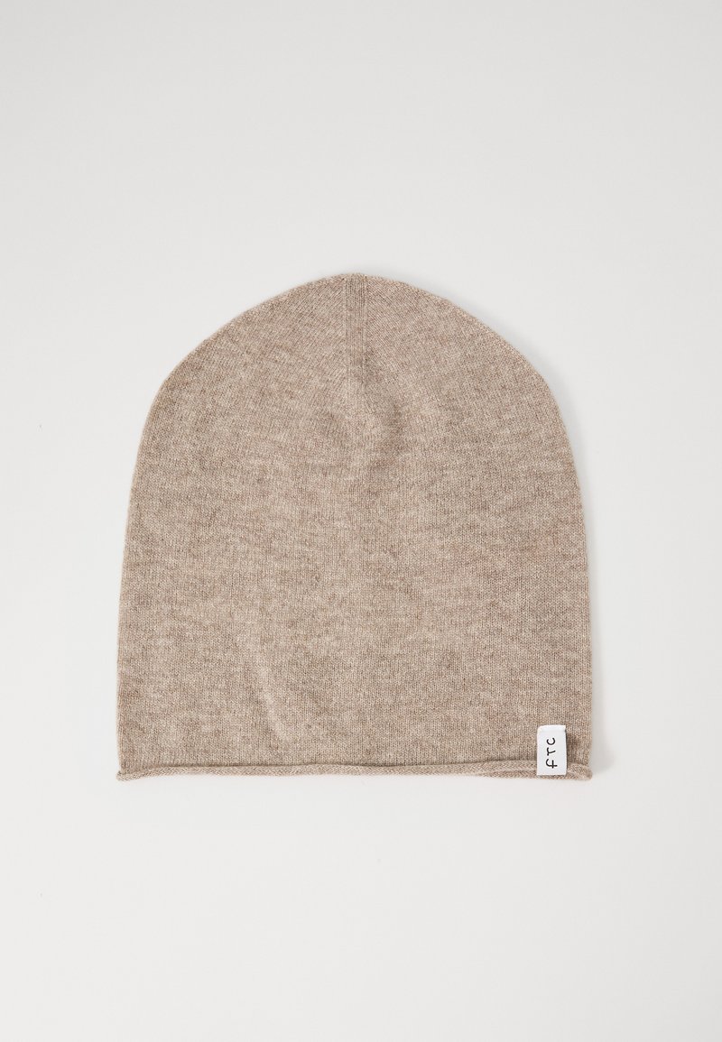 Beige knitted beanie with a small white fabric tag featuring black text on the bottom edge, laid flat on a white background.