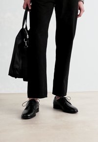Black leather shoes with a rounded toe, lace-up closure, and a low stacked heel. Accompanied by dark pants and a black bag.