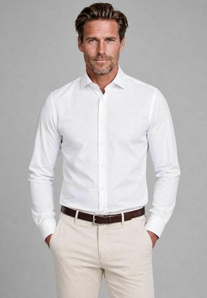 Man with styled brown hair wearing a white buttoned shirt, beige pants, and a dark brown belt with hands in pockets.