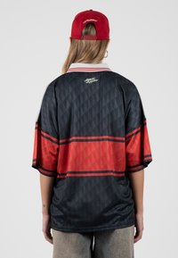 Persona con una camisa grande estampada en negro y rojo y gorra roja, de espaldas, de pie contra un fondo liso gris claro.