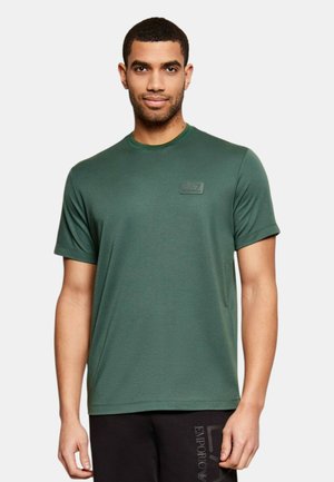 Uomo che indossa una T-shirt a maniche corte verde scuro con il logo EA7 sul petto e pantaloni neri Emporio Armani su sfondo uniforme.
