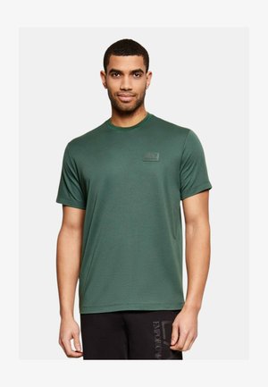 Uomo che indossa una T-shirt a maniche corte verde scuro con il logo EA7 sul petto e pantaloni neri Emporio Armani su sfondo uniforme.