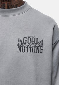Felpa grigia con collo rotondo, texture morbida e design stampato con farfalle. Il testo recita "PRESENTATA DA GOOD FOR NOTHING."
