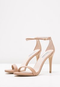 Ein Paar beiger High Heel Sandalen mit Knöchelriemen und offenen Zehen, auf einer weißen Fläche vor einem weißen Hintergrund präsentiert.